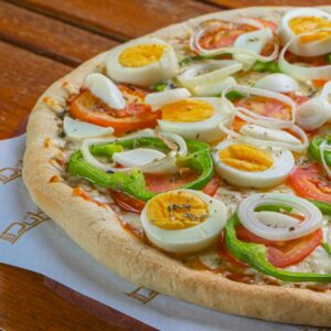Close-up of a pizza topped with eggs, onions, tomatoes, and peppers on a wooden table.