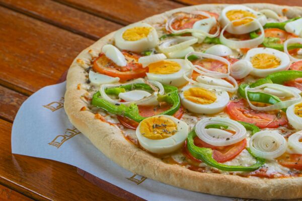 Close-up of a pizza topped with eggs, onions, tomatoes, and peppers on a wooden table.