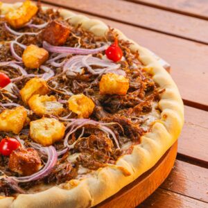 Close-up of a savory BBQ pulled pork pizza, garnished with red onions, croutons, and cherry tomatoes on a wooden board.